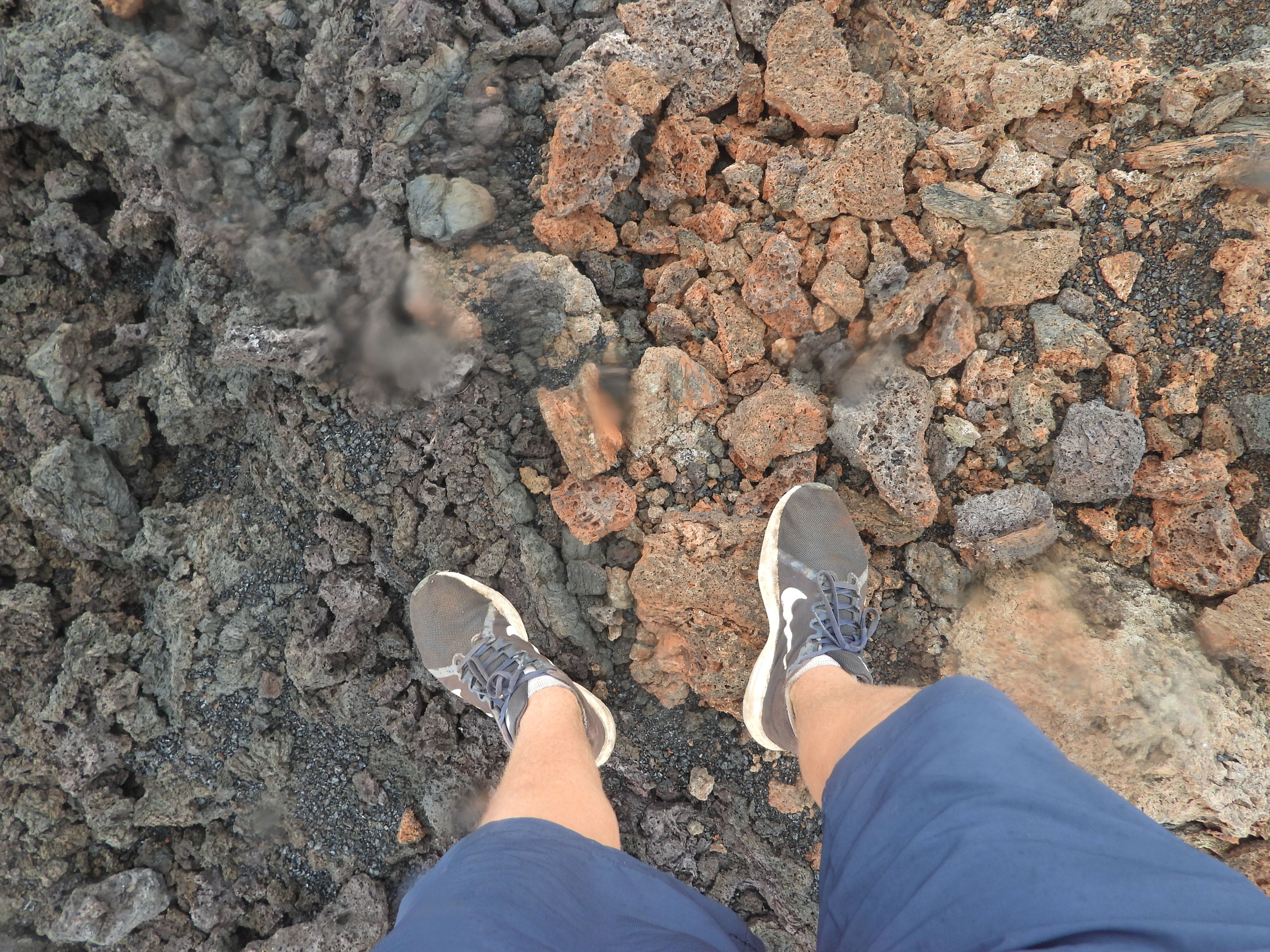 Feet standing on contrasting red and black basalt rocks at Sierra Negra Volcano, Galápagos, showing oxidation and cooling effects on volcanic lava through a rain-speckled lens.